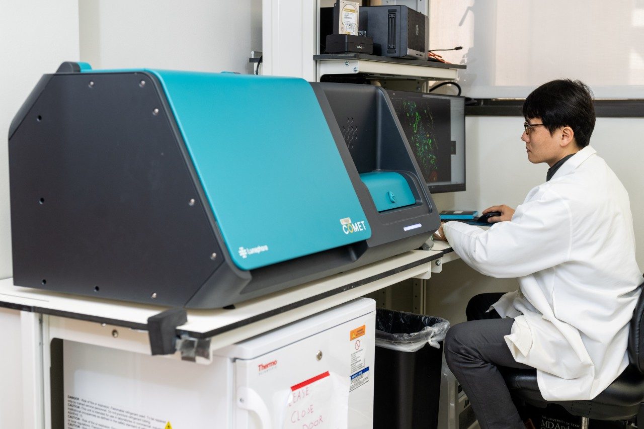 A person in a lab coat is seated at a workstation operating a computer connected to a large teal-colored instrument labeled “COMET.” The screen shows scientific data or images, and lab equipment is visible on surrounding shelves.