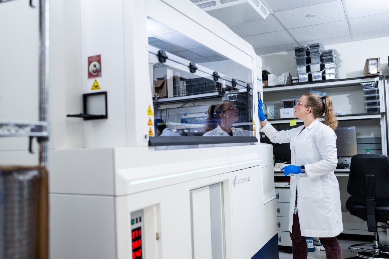 A person in a lab coat and gloves is interacting with a large enclosed laboratory instrument. The machine has a transparent front panel and occupies most of the foreground. Shelves with lab supplies are visible in the background.