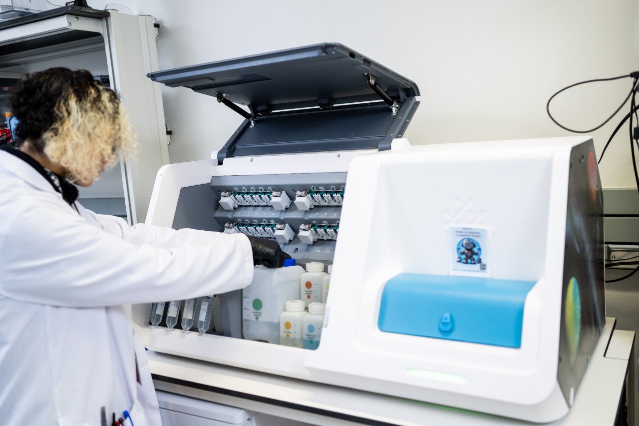A person in a lab coat is loading or arranging bottles inside a large white instrument with an open lid. The machine has multiple slots for containers and a blue compartment on the right side.