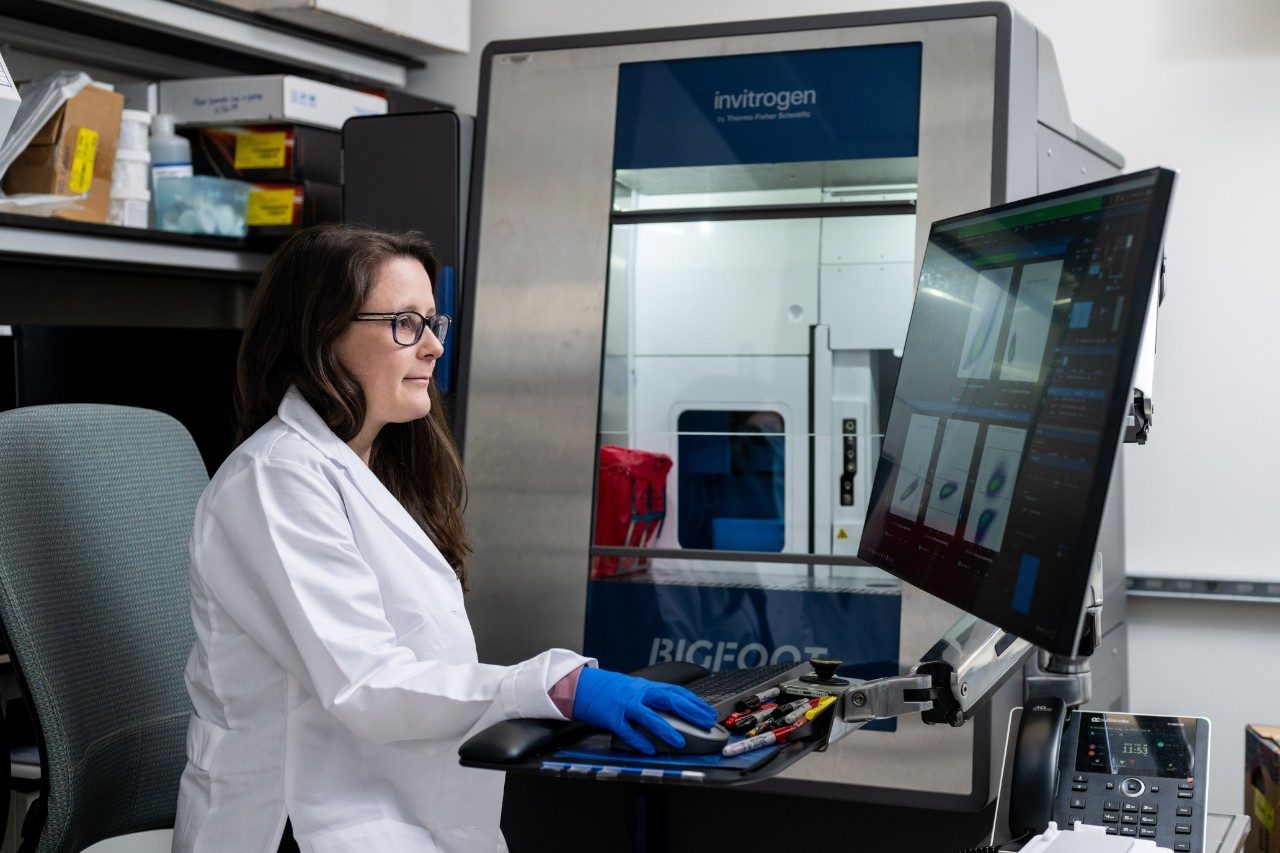 A person in a lab coat is seated at a workstation operating a computer connected to an Invitrogen Bigfoot instrument. The computer screen displays graphical data, and the instrument is positioned behind the monitor.
