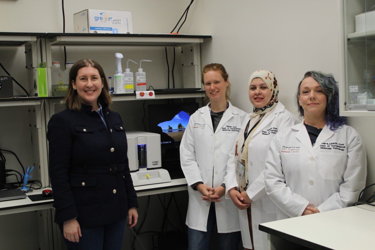 ORION group photo. Four individuals standing in a laboratory next to a benchtop instrument and a computer monitor displaying a blue screen. Shelves above the bench hold bottles, pipettes, and other lab supplies.