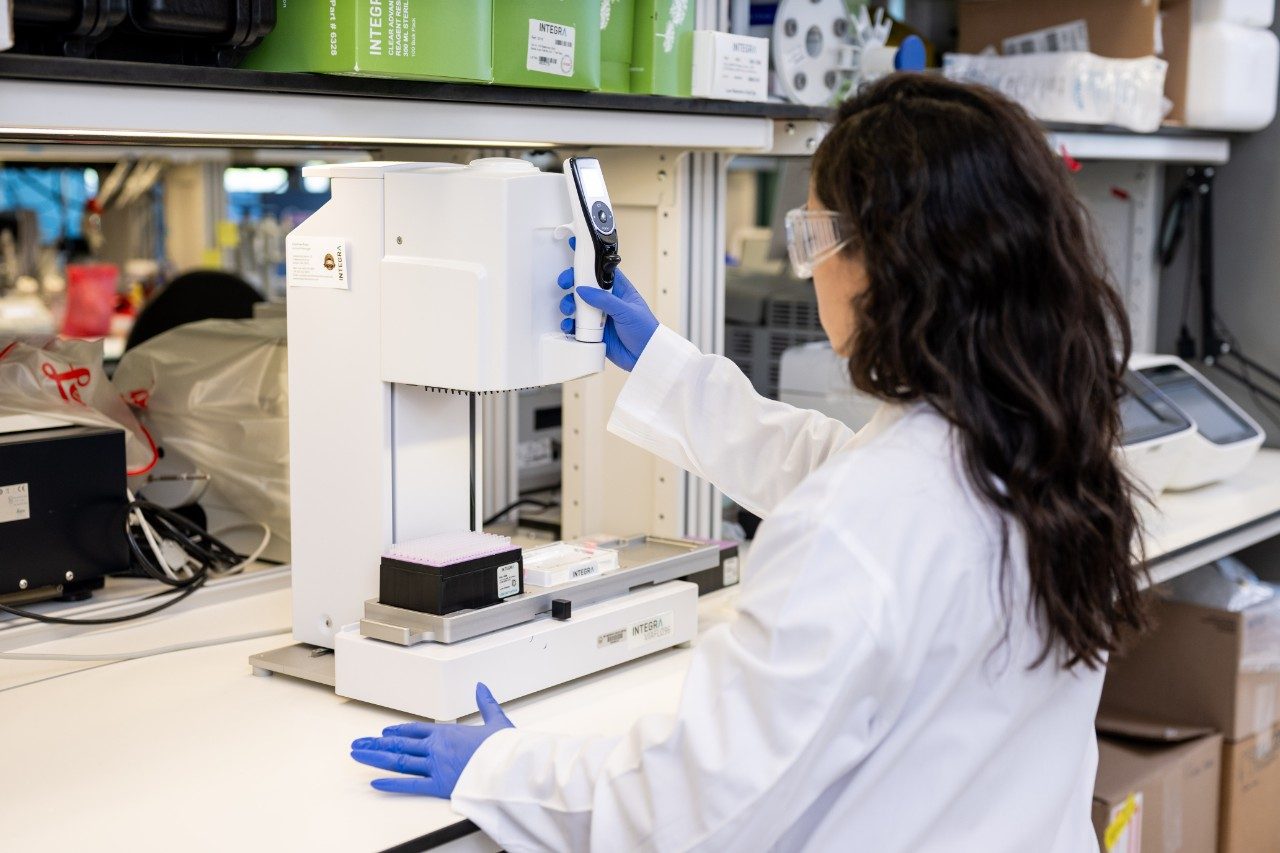 A person in a white lab coat and blue gloves is operating a benchtop instrument with a vertical component and a tray of samples. The individual is holding a handheld device near the machine. Shelves with boxes and other lab equipment are visible in the background.