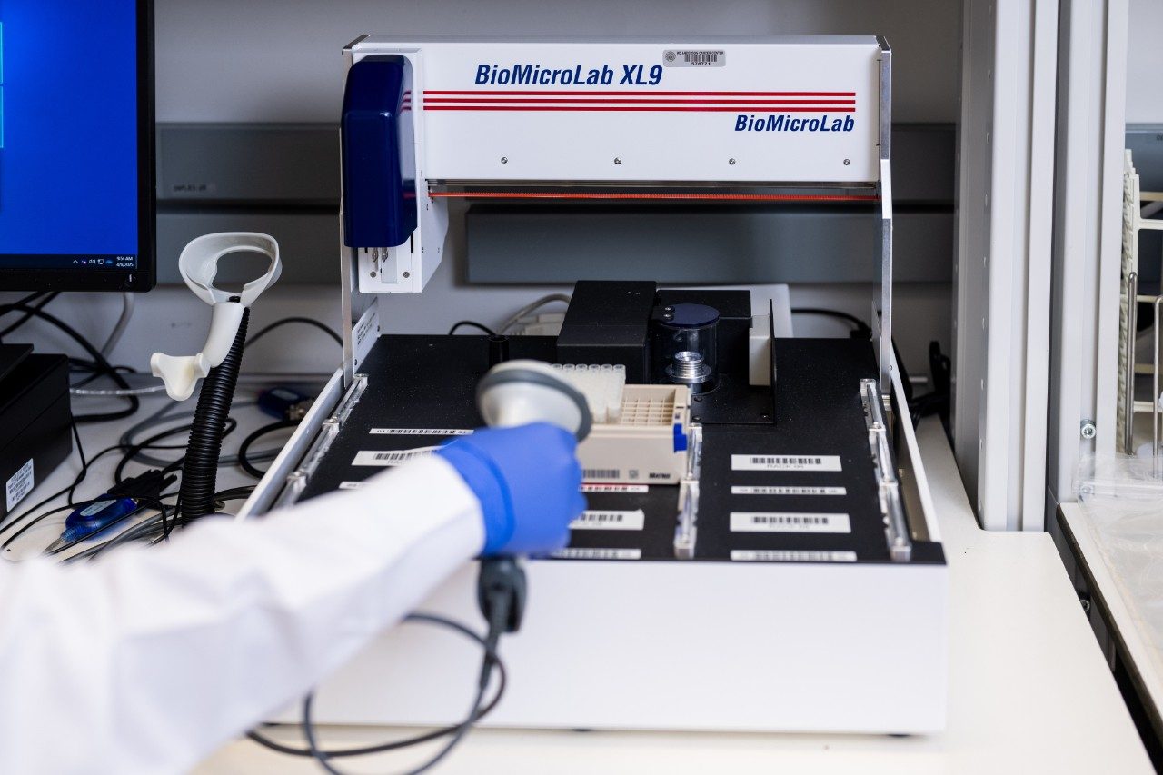 Close-up of a BioMicroLab XL9 instrument on a laboratory bench. A gloved hand is holding a barcode scanner near a rack of labeled sample tubes inside the machine. A computer monitor and other lab equipment are visible in the background.