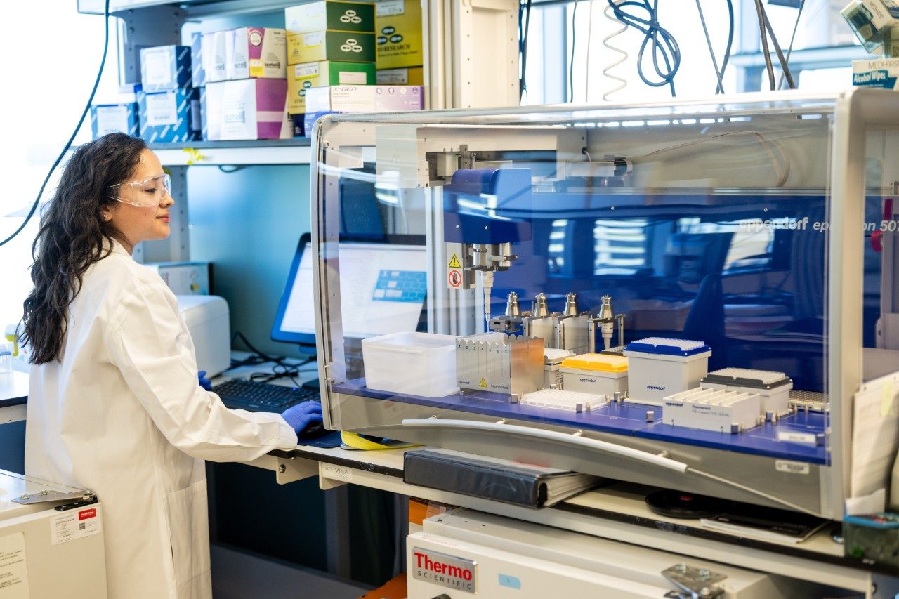 A person in a white lab coat is standing in front of an automated liquid handling instrument inside a clear enclosure. The machine contains multiple racks of pipette tips, sample trays, and containers. A computer monitor displaying data is positioned behind the instrument, and shelves with colorful boxes of lab supplies are visible above.