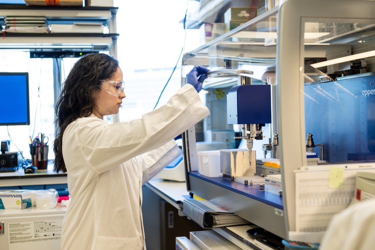 A person wearing a white lab coat and blue gloves is working at a laboratory bench with an automated liquid handling system inside a protective enclosure. The individual is adjusting the lid of the machine, which contains pipette tips and sample trays. Shelves with lab supplies and a computer monitor are visible in the background.