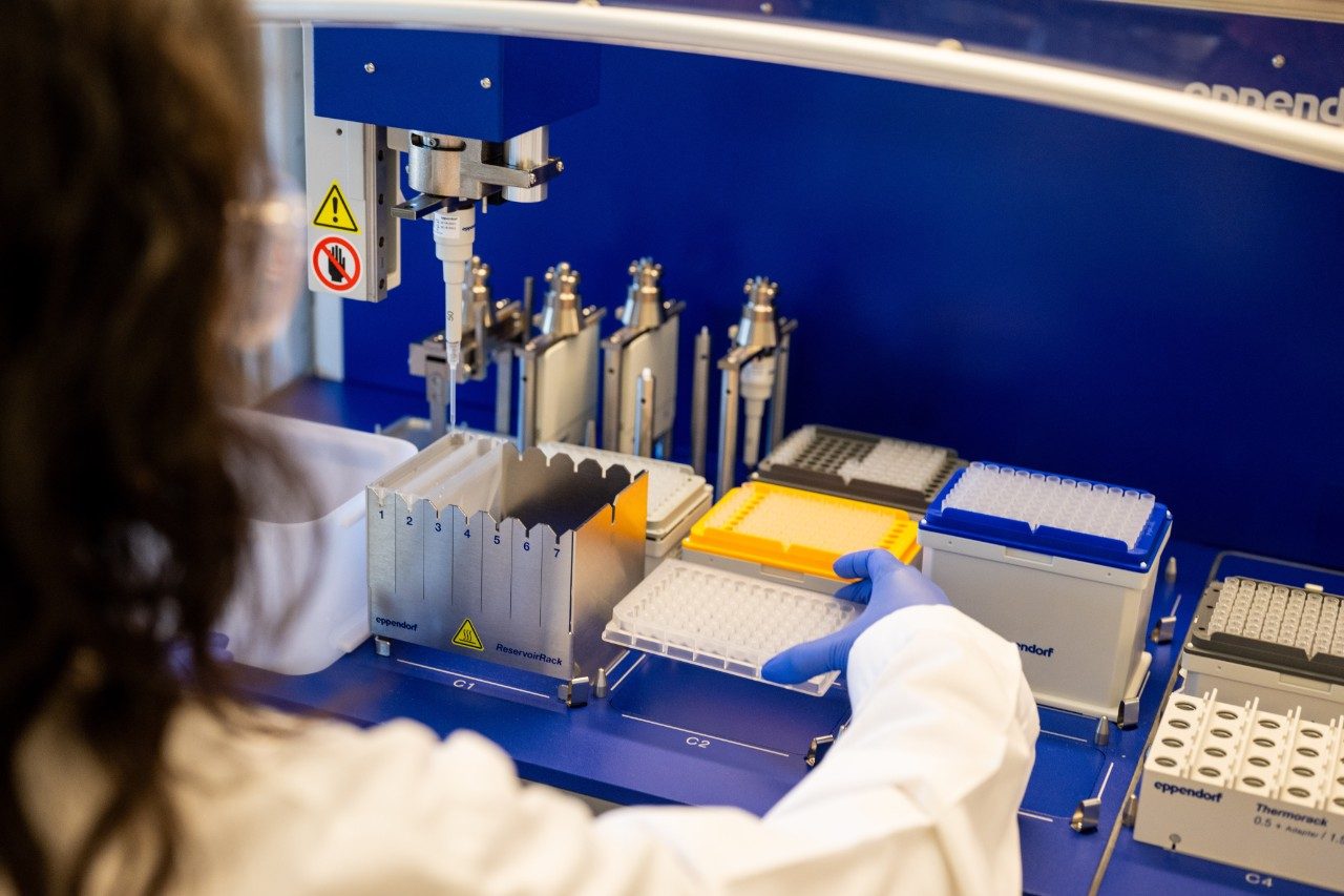 Three individuals standing in a laboratory between two rows of benches. Two people are wearing white lab coats with institutional logos, and the third person is wearing a white button-down shirt. Laboratory equipment, shelves with supplies, and a large window showing buildings outside are visible in the background.