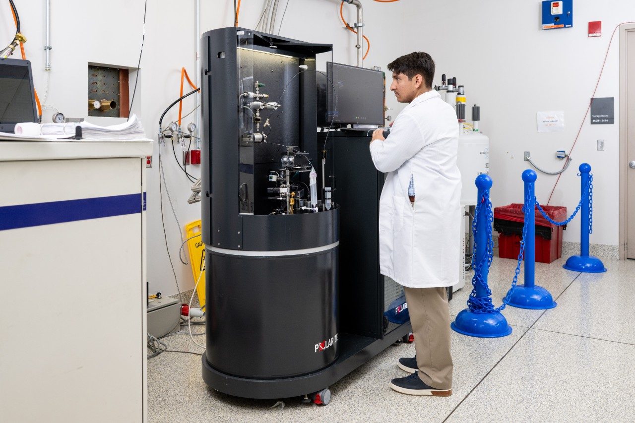 A person in a white lab coat is operating a large black instrument labeled “Polaris” in a laboratory. The machine has an open compartment with visible components and a mounted monitor. The surrounding area includes electrical cables on the wall and blue stanchions in the background.