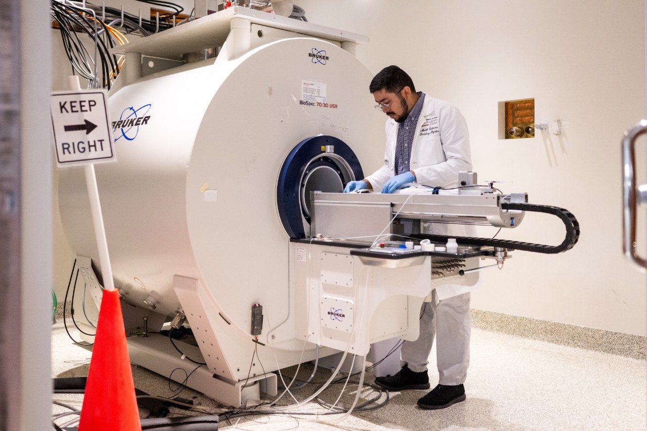 A person wearing a white lab coat and blue gloves is working with a large cylindrical imaging machine labeled “Bruker.” The machine has multiple cables connected to it and a flat platform extending outward. A sign reading “Keep Right” and an orange traffic cone are visible nearby.