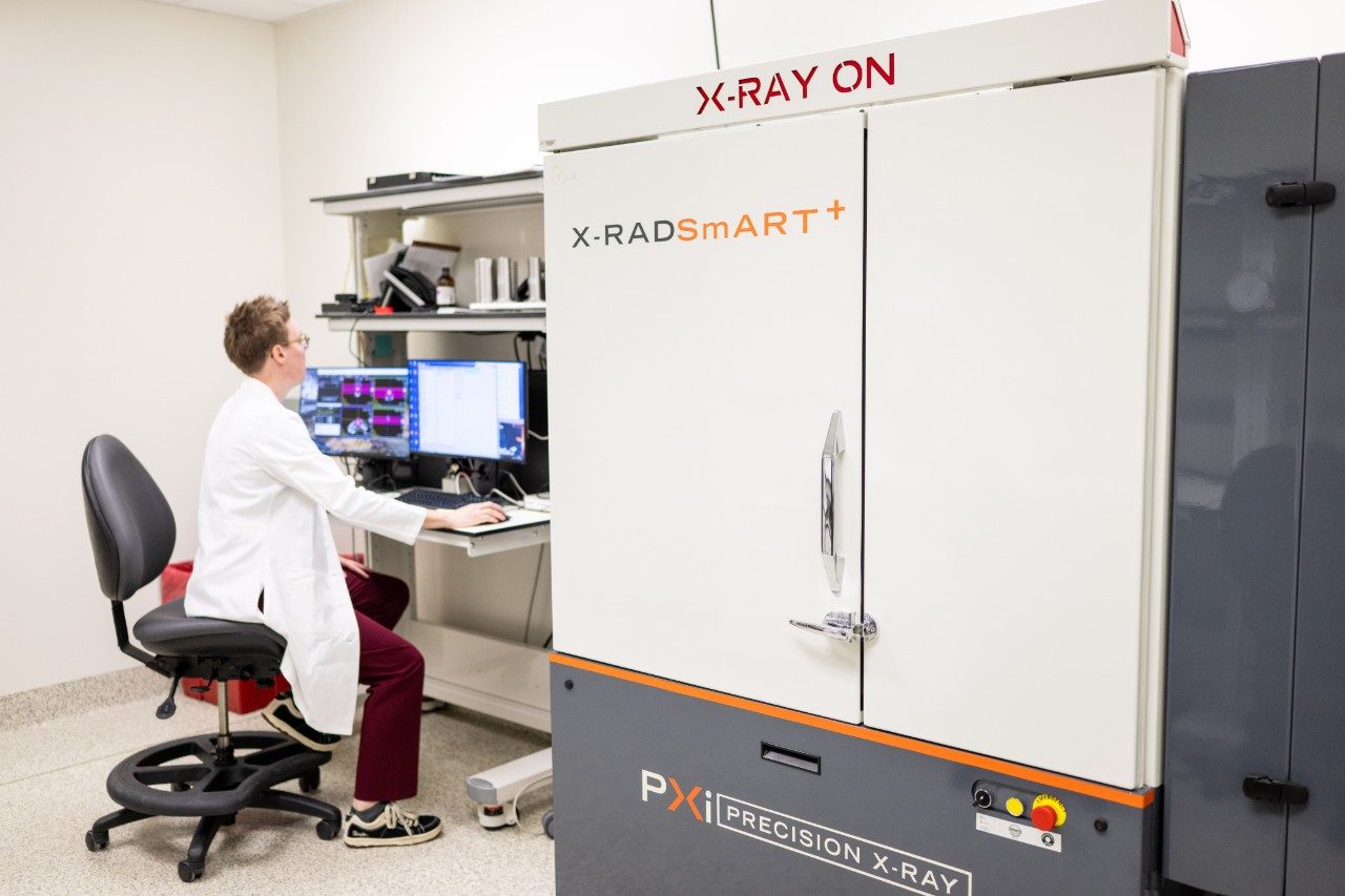 A person in a white lab coat is seated at a workstation operating a computer connected to an X-ray imaging system labeled “X-RADs mART+” by PXi Precision X-Ray. The machine has a large closed compartment with “X-RAY ON” displayed in red at the top. The computer monitors show imaging software and data.