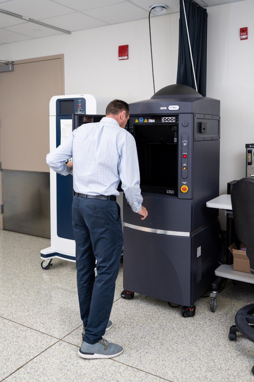 A person is standing in front of a tall black instrument labeled “iCOOL” in a laboratory. The machine has an open compartment and control buttons on the front panel. Another piece of equipment on wheels is positioned nearby, and a desk with a chair is visible in the background.