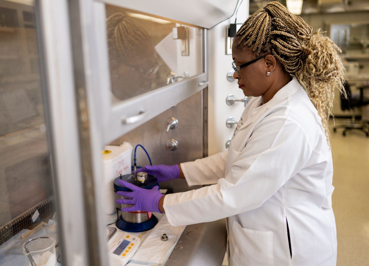 A RHCL staff member in a white coat and purple gloves operates equipment inside a biosafety cabinet.