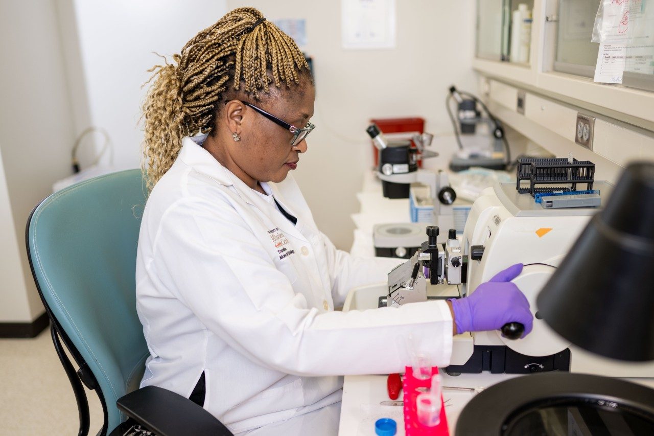 A RHCL staff member in a white coat and purple gloves operates a microtome at a laboratory bench.