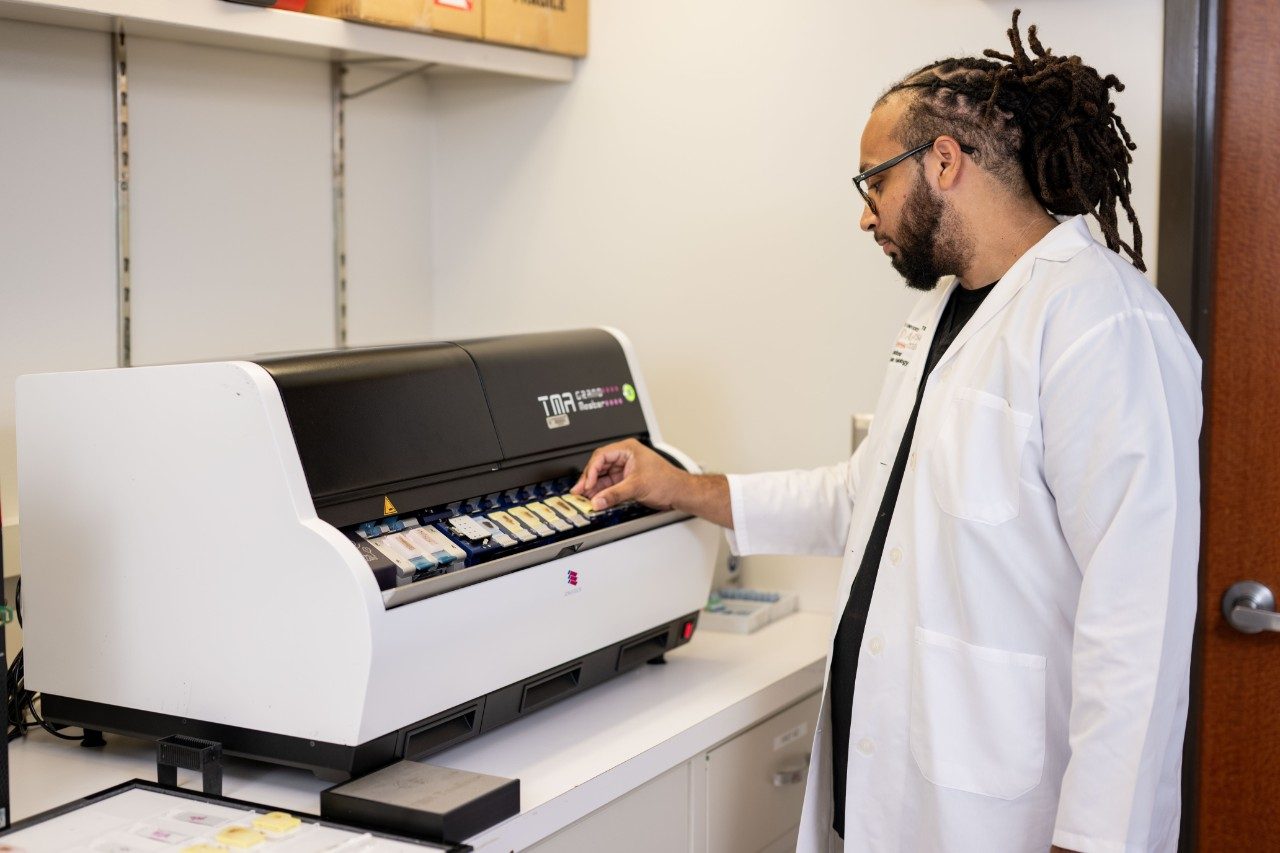 A RHCL staff member in a white coat operates a slide staining machine on a countertop in a laboratory.